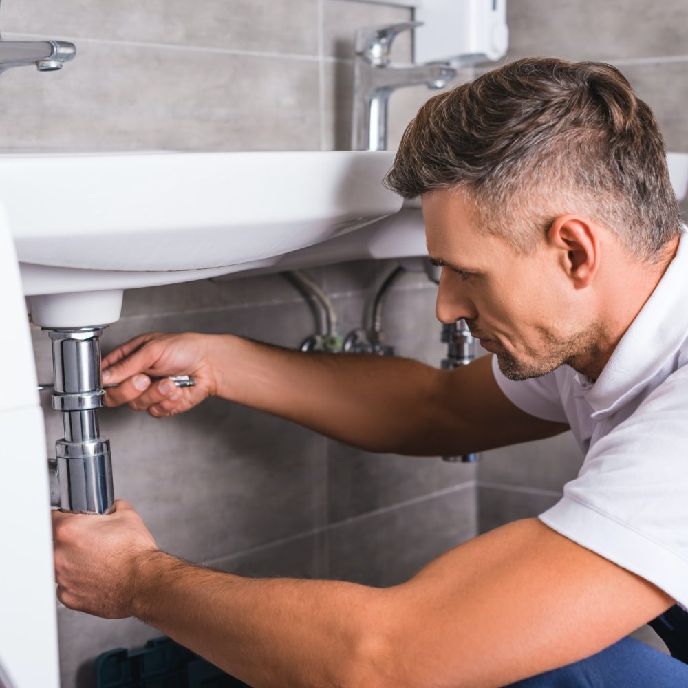 A plumber in a white shirt and overalls works under a sink, tightening pipes with a wrench. The setting is a modern bathroom with gray tiles, conveying focus.