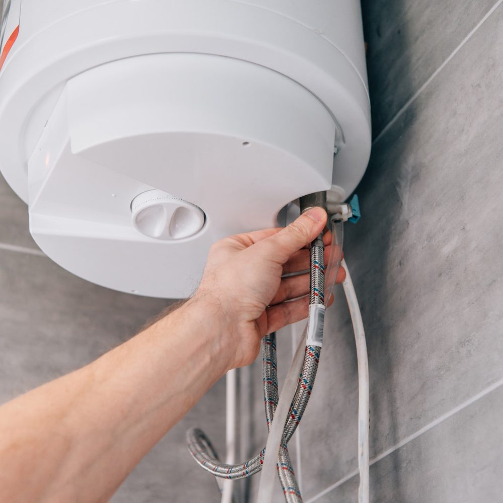 A person adjusts the connection of a water heater in a tile-walled room. The hand is securing braided hoses to the white appliance, focusing on plumbing work.