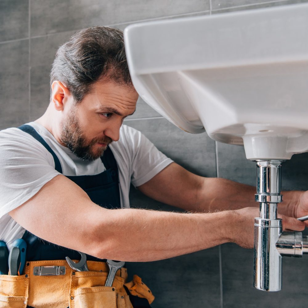 A focused plumber in a white shirt and overalls is repairing a sink in a modern bathroom. He uses a wrench, tools visible in his yellow tool belt.