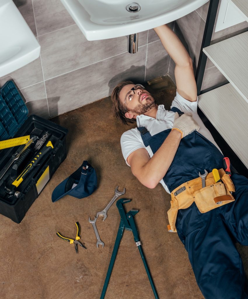 A plumber in blue overalls lies on a tiled floor, fixing a sink. Nearby, a toolbox and various tools are scattered. The scene conveys focus and diligence.