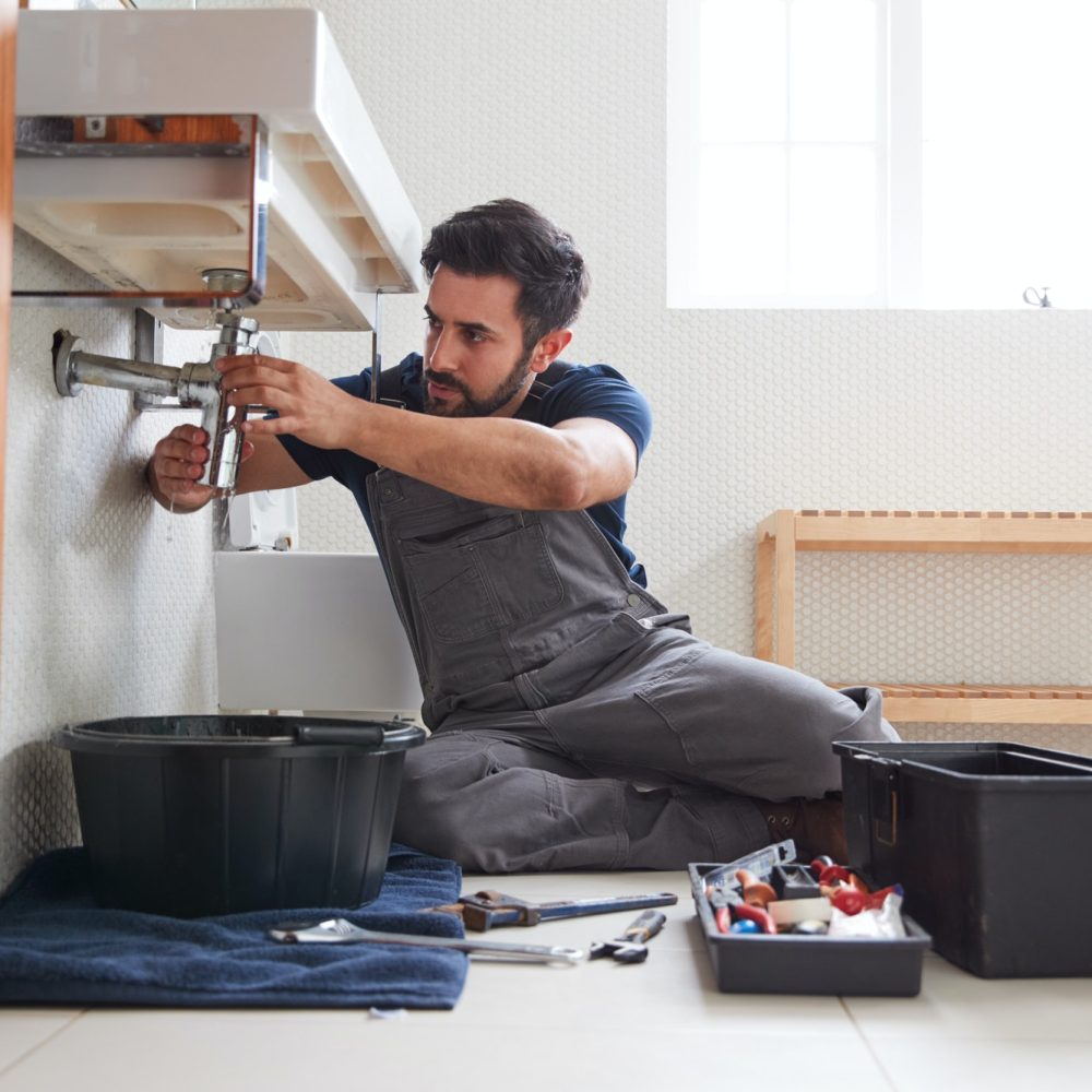 A plumber in overalls is fixing pipes under a sink in a well-lit bathroom, with tools and a black toolbox nearby, conveying focus and professionalism.