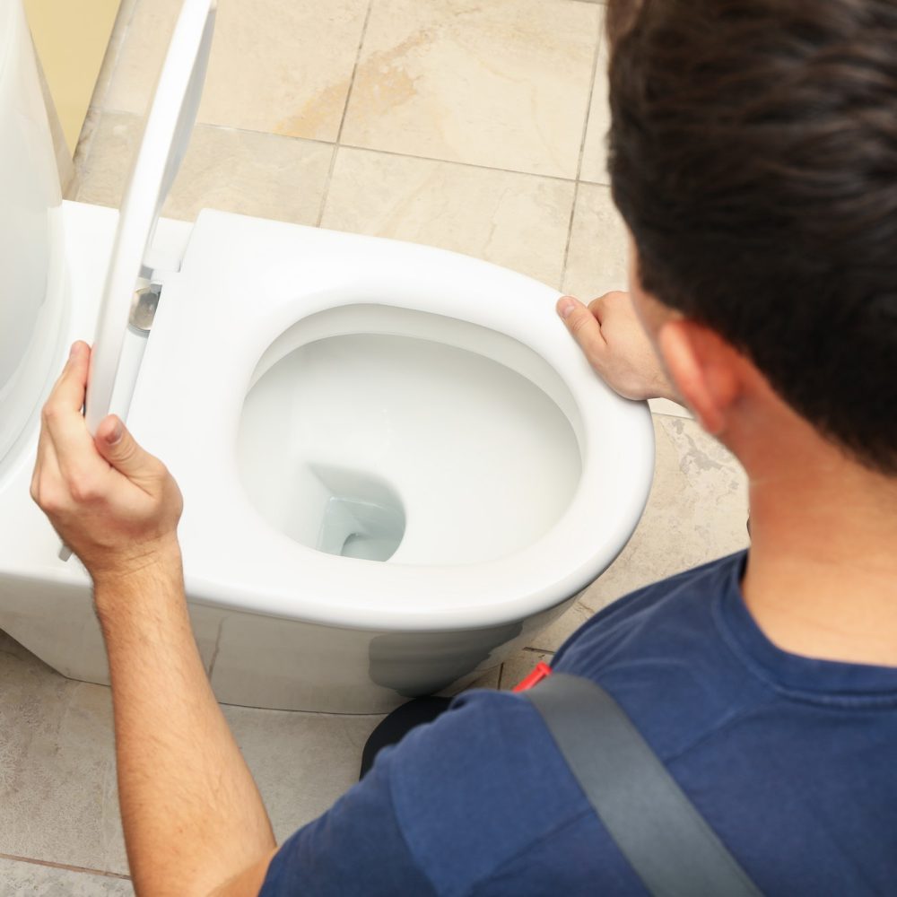A person in a blue shirt lifts a toilet lid, peering inside. The bathroom has beige tiled flooring, and the scene suggests maintenance or inspection.
