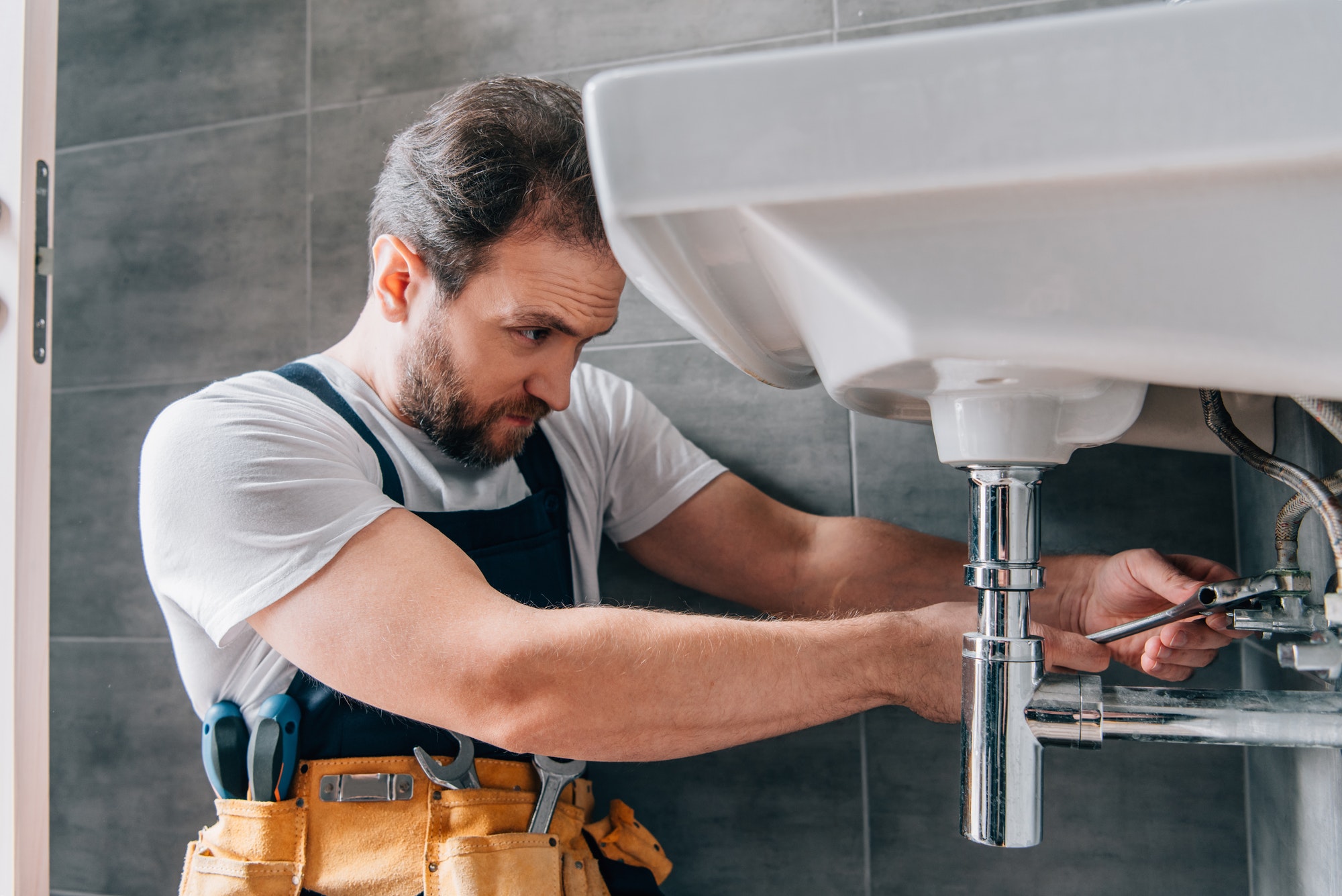 A focused plumber in a white shirt and overalls is repairing a sink in a modern bathroom. He uses a wrench, tools visible in his yellow tool belt.